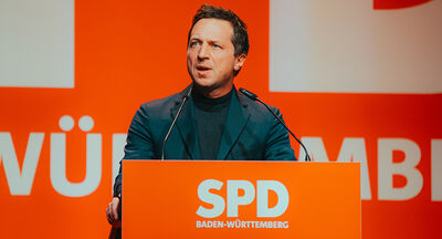 Oberkörperaufnahme von Sascha Binder, Generalsekretär der SPD Baden-Württemberg, auf einer Bühne sprechend. Am Rednerpult weiß auf rot das Logo der SPD Baden-Württemberg. Ebenfalls weiß auf rot ist das Logo auch im Hintergrund zu sehen.